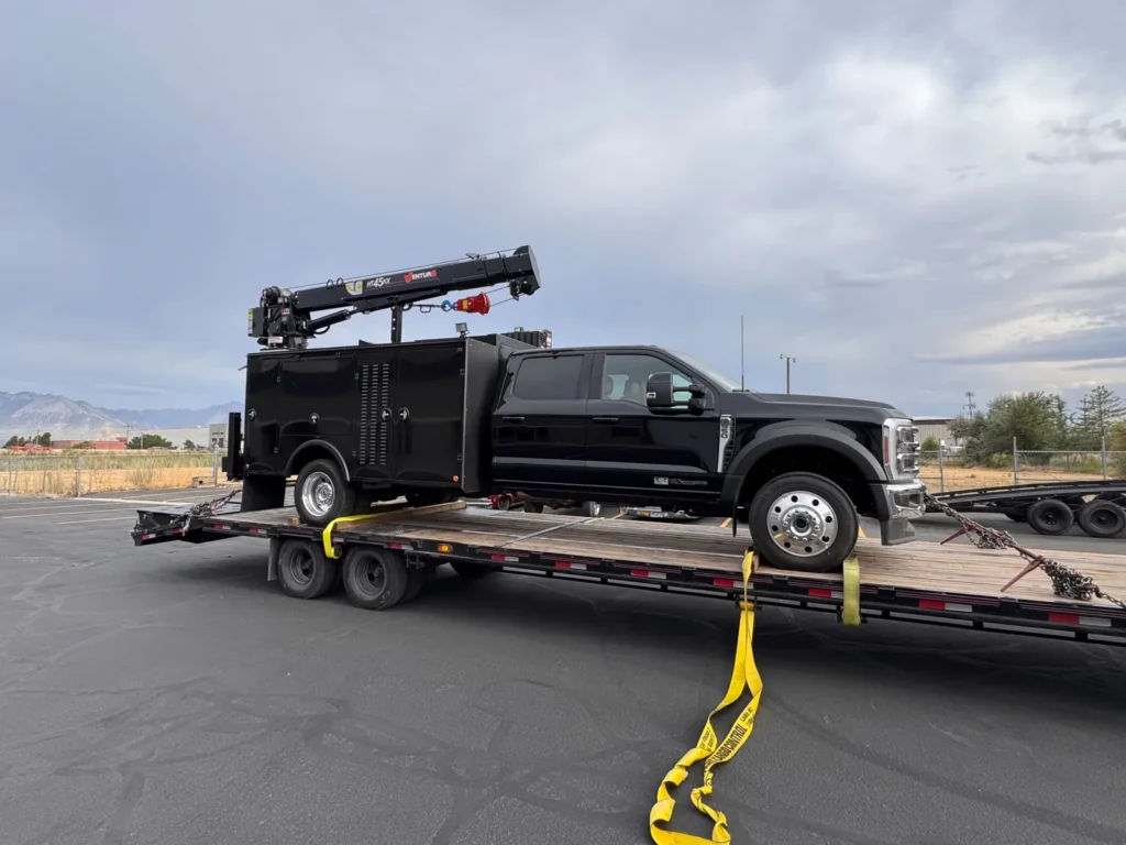Black service-body truck with mounted crane positioned on a flatbed trailer